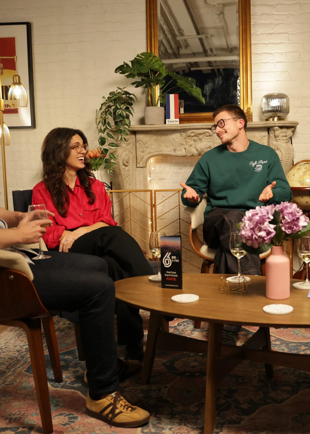 Four people are seated in a cozy, warmly lit living room around a wooden coffee table, engaging in conversation. The room features a white brick fireplace, a gold-framed mirror, and potted plants in the background.