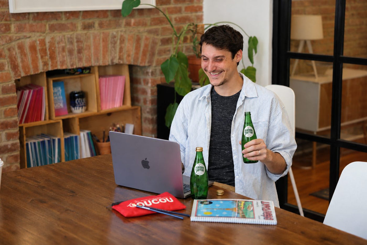 A Coucou student smiling at his laptop with two green glass bottles of Maison Perrier® and a Coucou notebook on the table