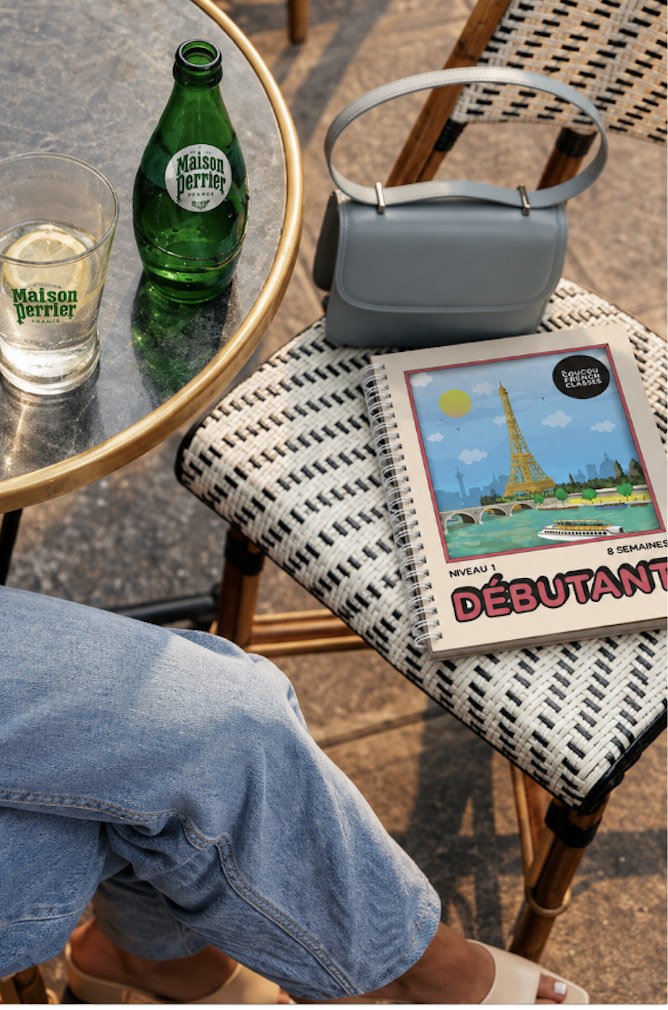 A Paris terrasse table with a green glass bottle of Maison Perrier®, a glass with lemon and a Coucou Débutant notebook on a bistro chair